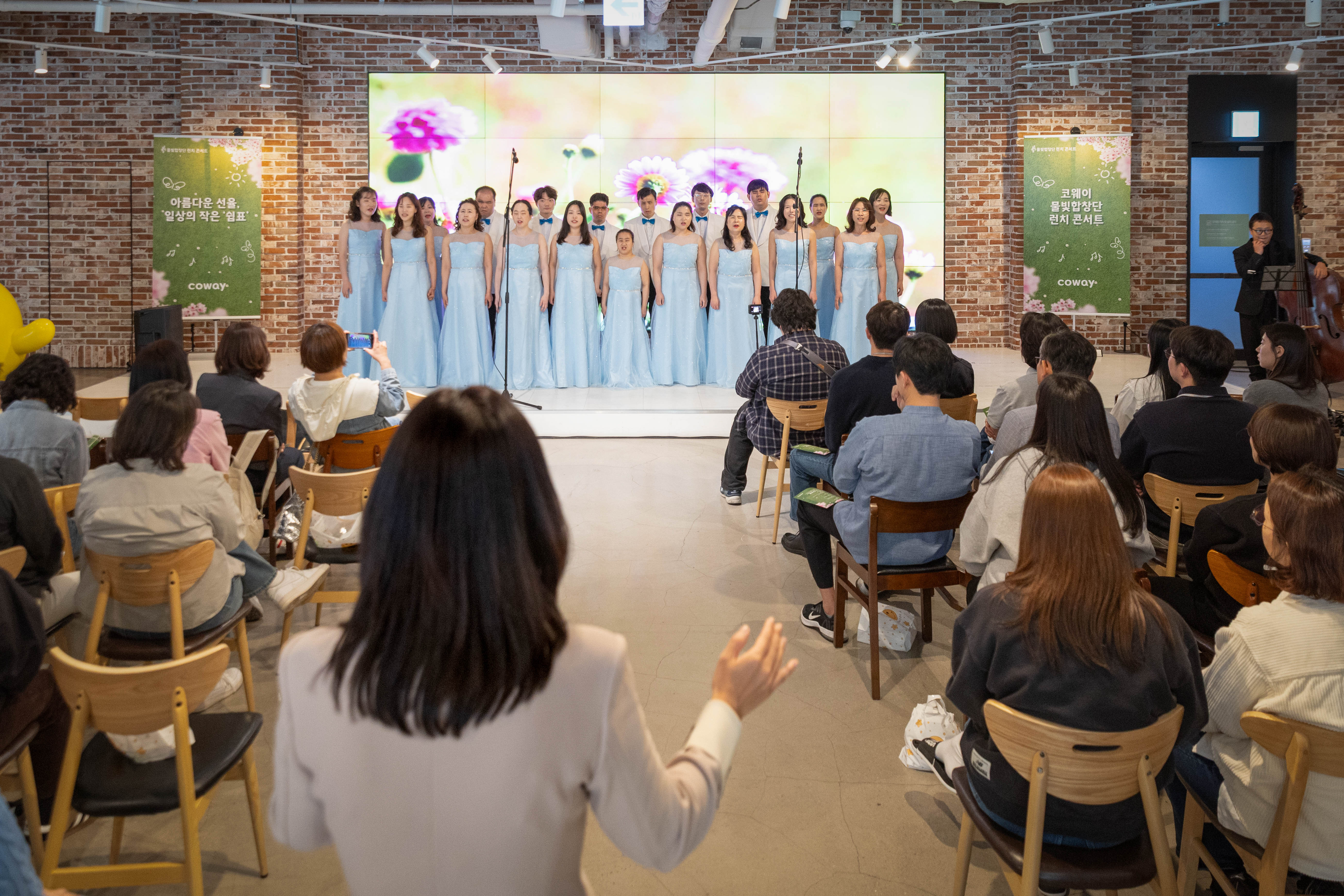 2026년 4월 16 서울 구로구 지타워 내 위치한 'ㅋㅋ다방'에서 코웨이 물빛합창단이 제4회 런치콘서트를 진행했다. 사진 중앙에는 함정민 지휘자가 지휘를 하고 있으며, 무대에는 물빛합창단 단원들이 합창 무대를 선보이고 있다. 무대 앞은 관객들로 가득 차 있다.