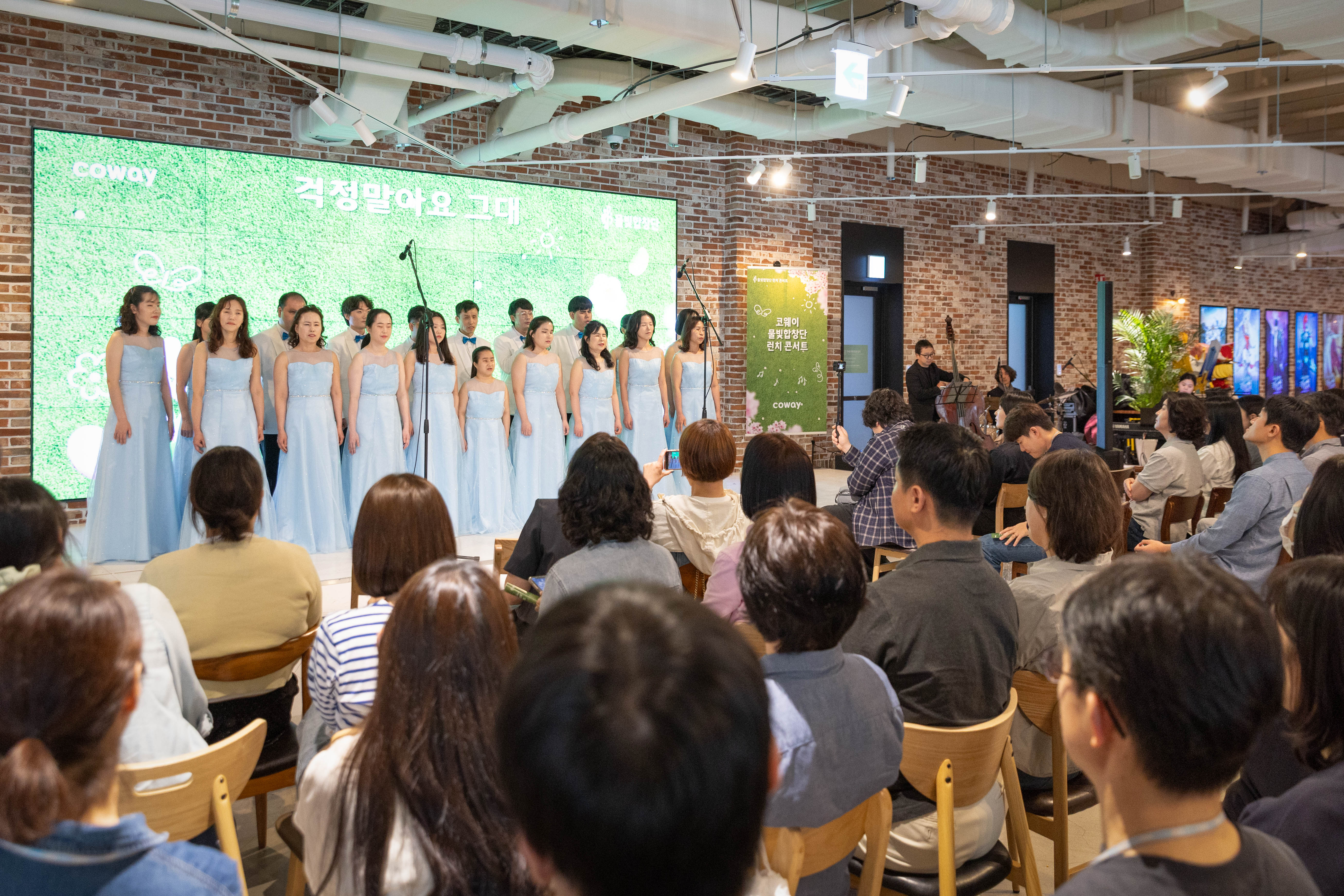 2026년 4월 16 서울 구로구 지타워 내 위치한 'ㅋㅋ다방'에서 코웨이 물빛합창단이 제4회 런치콘서트를 진행했다. 무대에는 물빛합창단 단원들이 합창 무대를 선보이고 있다. 무대 옆에는 '블루 브리즈 콰르텟'이 연주를 선보이고 있으며, 무대 앞은 관객들로 가득 차 있다.