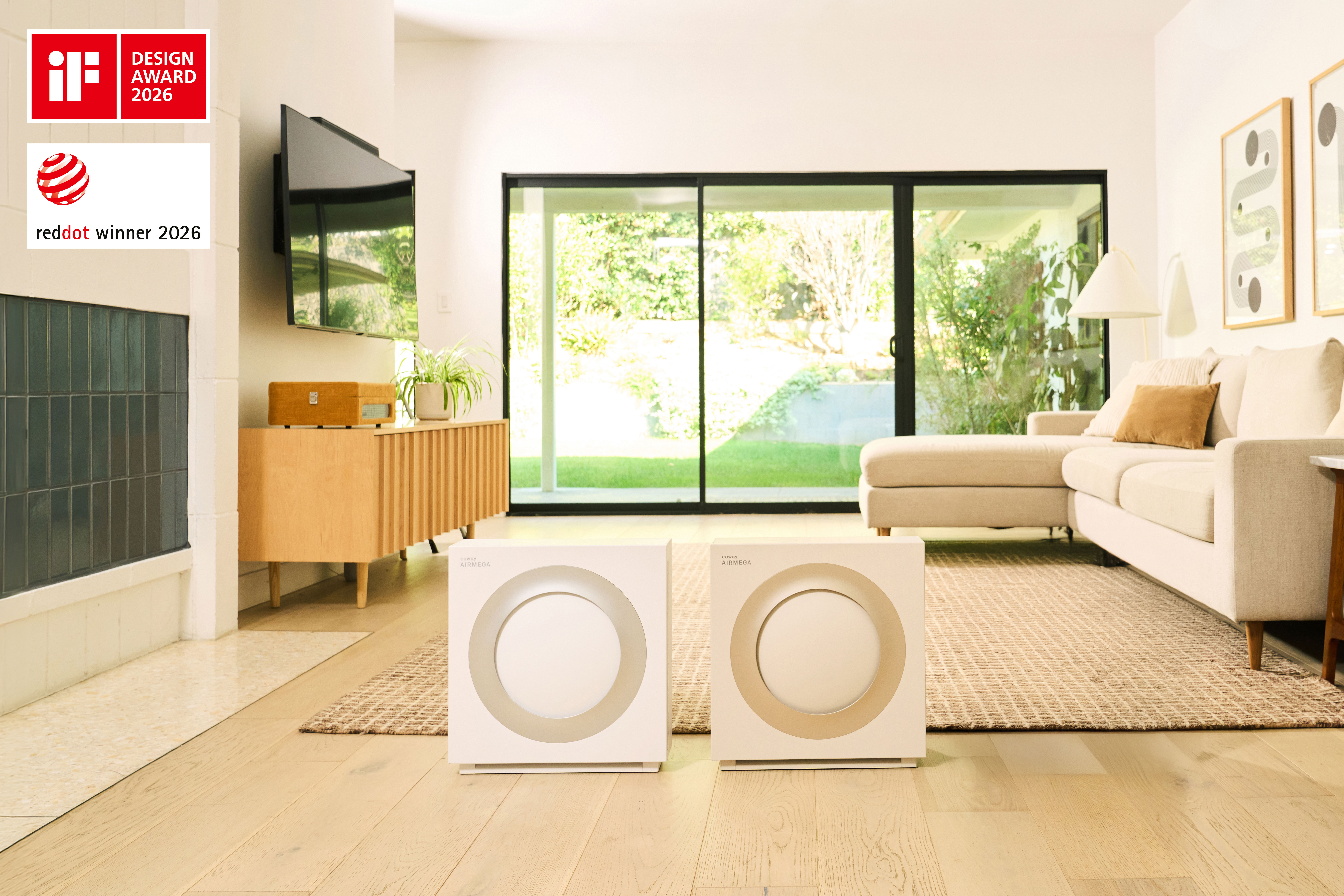 Two square-shaped air purifiers, the Airmega Mighty2, are placed side-by-side on a light wood floor in a sunlit modern living room. The unit on the left features a light grey circular accent, while the one on the right has a warm beige circular accent. The background includes a stylish wooden console, a record player, a cream-colored sectional sofa, and large glass sliding doors looking out to a garden.