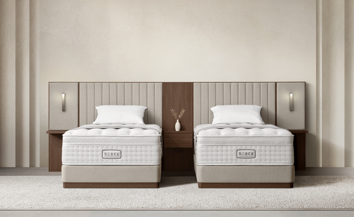 뉴트럴 베이지 컬러의 비렉스 스위트 호텔 프레임에 중앙 우드 패널과 양옆 패브릭 패널을 조합한 '트리플 패널' 구성 사진입니다. 중앙의 월넛 우드 패널 서랍 위에는 흰색 꽃병과 나뭇가지 소품이 놓여 있고, 양옆의 멀티 패널에는 무드 조명이 켜져 있어 따뜻한 분위기를 연출합니다. 파운데이션 하부는 목재 몰딩으로 마감되었으며, 투매트리스 구조와 E0 등급 자재를 사용하여 안정성과 안전성을 높인 프리미엄 침대입니다.